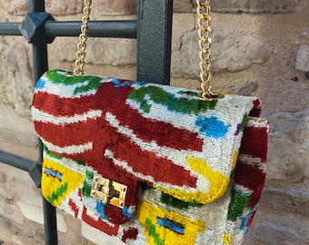 May include: A small, colorful, patterned handbag with a gold chain strap. The bag is made of a soft, velvety fabric and has a flap closure with a gold clasp.