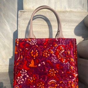 May include: A red, orange, and purple floral patterned tote bag with a light brown leather handle.
