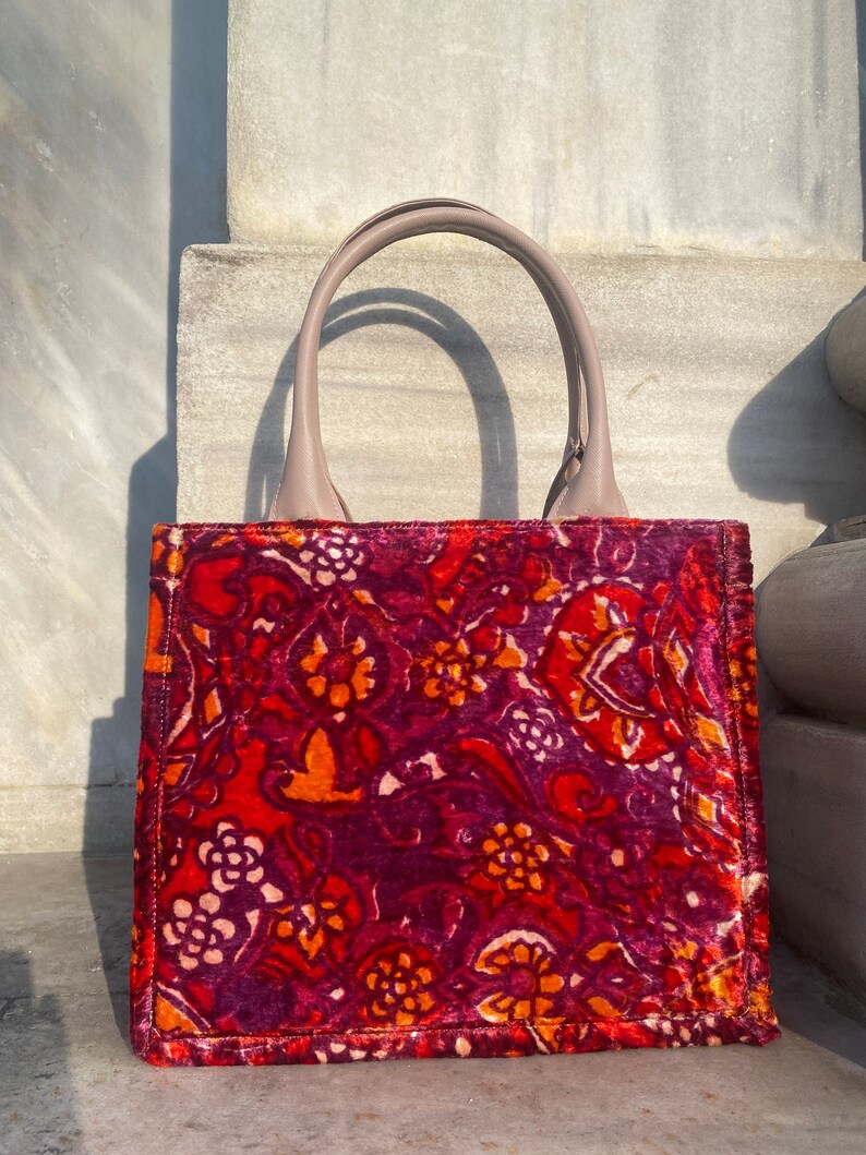 May include: A red, orange, and purple floral patterned tote bag with a light brown leather handle.