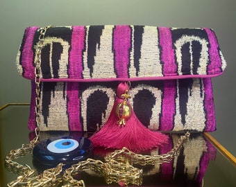 May include: A pink and black patterned clutch bag with a gold chain strap and a pink tassel with a gold pomegranate charm. The bag is on a reflective surface with a blue evil eye charm.