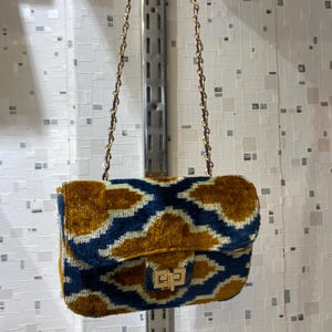 May include: A small, rectangular handbag with a gold chain strap. The bag is made of a plush, patterned fabric in shades of navy blue and gold. It features a gold clasp closure.