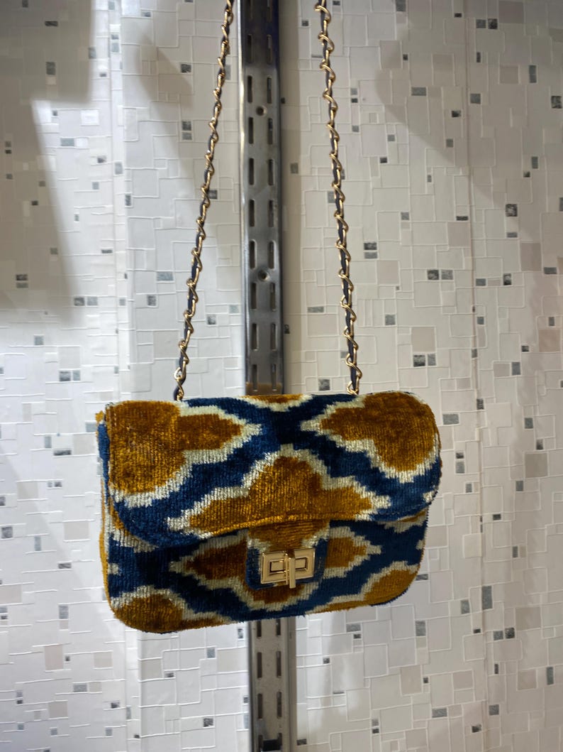 May include: A small, rectangular handbag with a gold chain strap. The bag is made of a plush, patterned fabric in shades of navy blue and gold. It features a gold clasp closure.