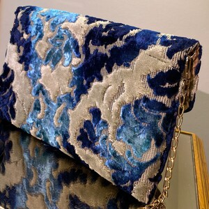 Op de afbeelding: Een blauwe en grijze clutch met patroon en een gouden kettingband. De tas heeft een bloemenmotief en is gemaakt van een zachte, gestructureerde stof.
