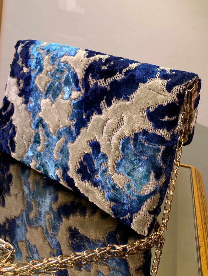 Op de afbeelding: Een blauwe en grijze clutch met patroon en een gouden kettingband. De tas heeft een bloemenmotief en is gemaakt van een zachte, gestructureerde stof.