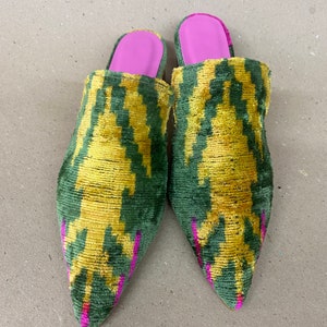 May include: A pair of pointed-toe mules with a green and yellow patterned design. The shoes have a pink interior and are made of a textured fabric. The mules are a slip-on style.