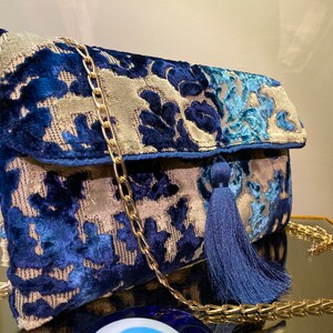 Op de afbeelding: Een blauwe en beige velvet clutch met een gouden kettingband en een blauwe kwast. De tas heeft een bloemenpatroon.