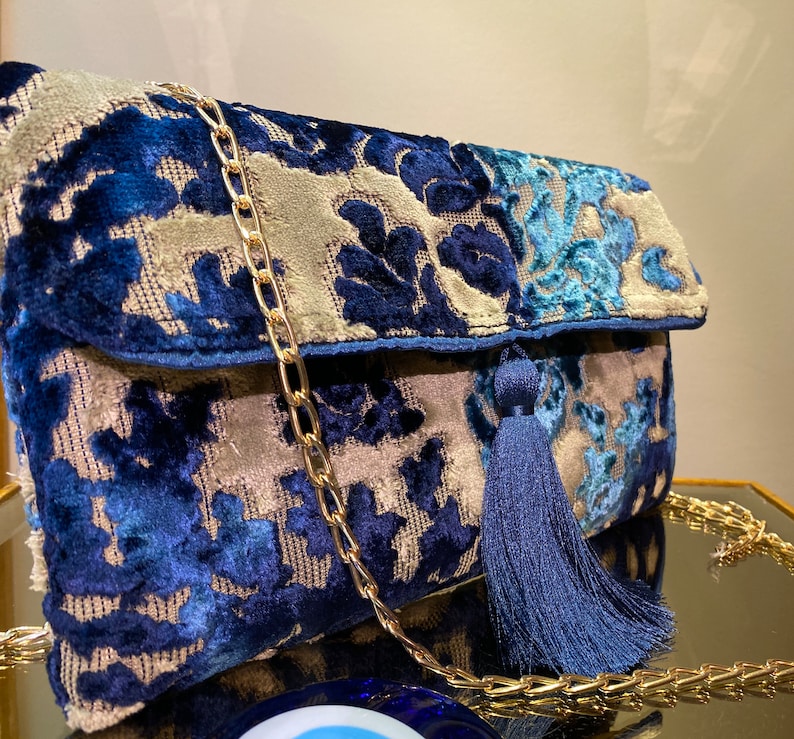 Op de afbeelding: Een blauwe en beige velvet clutch met een gouden kettingband en een blauwe kwast. De tas heeft een bloemenpatroon.