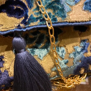 Op de afbeelding: Een blauwe en gouden stoffen clutch met een kwast en een gouden kettingband.