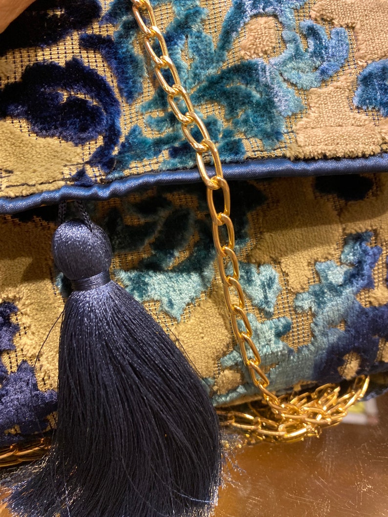 Op de afbeelding: Een blauwe en gouden stoffen clutch met een kwast en een gouden kettingband.