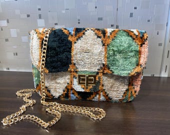 May include: A rectangular handbag with a gold chain strap. The bag features a colorful, patterned fabric in shades of green, black, orange, and blue. The clasp is gold-toned.