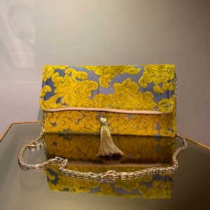 K&ouml;nnte beinhalten: Eine goldene und graue Clutch mit Quaste. Die Clutch hat einen Kettenriemen und ein Blumenmuster.