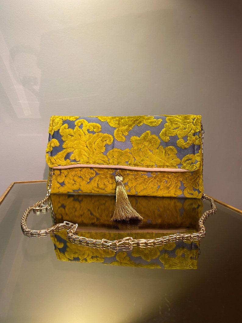 K&ouml;nnte beinhalten: Eine goldene und graue Clutch mit Quaste. Die Clutch hat einen Kettenriemen und ein Blumenmuster.