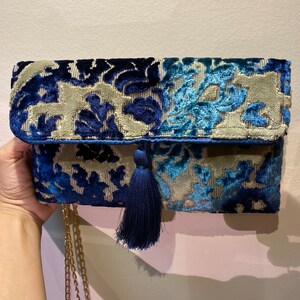 Op de afbeelding: Een blauwe en beige fluwelen clutch met een kwast en een gouden kettingband. De clutch heeft een bloemenpatroon.