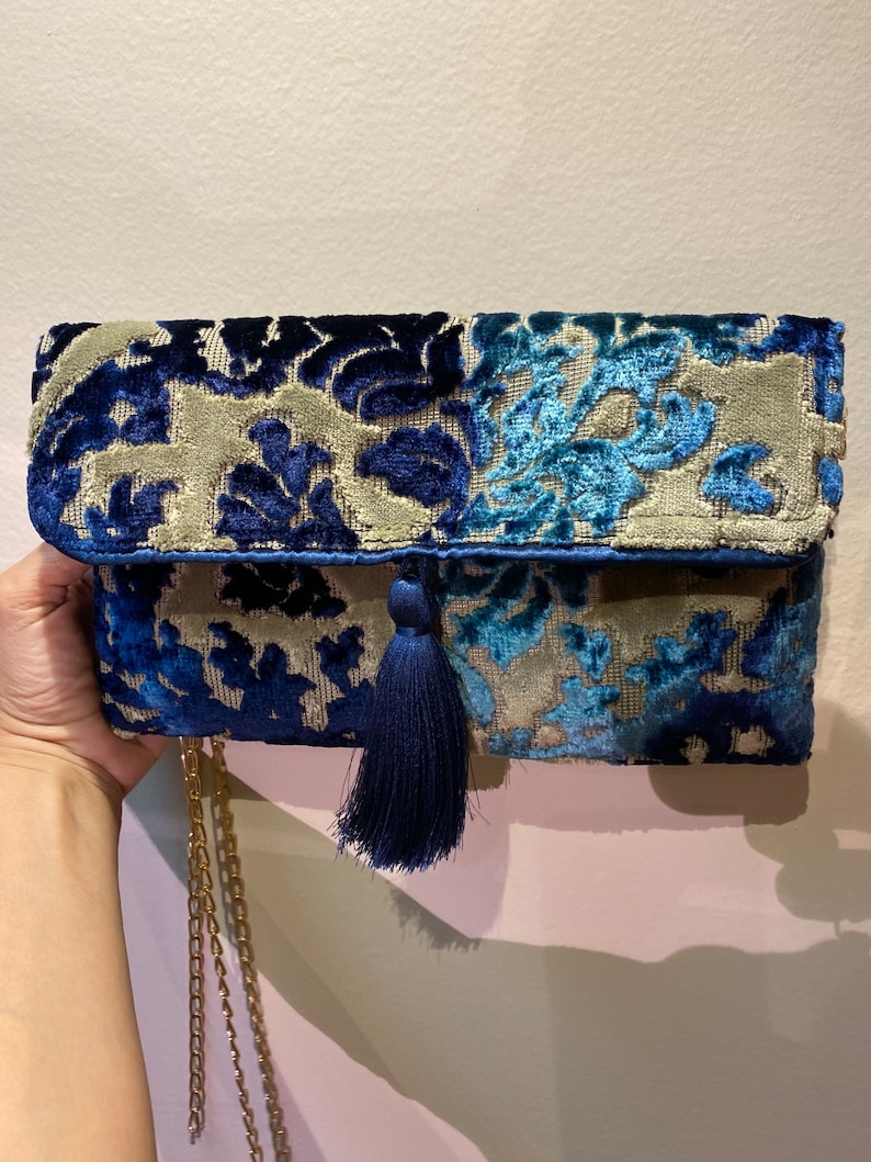 Op de afbeelding: Een blauwe en beige fluwelen clutch met een kwast en een gouden kettingband. De clutch heeft een bloemenpatroon.