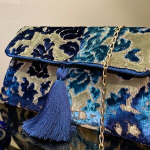 Op de afbeelding: Een blauwe en grijze velvet clutch met een gouden kettingband en een blauwe kwast. De tas heeft een bloemenpatroon.