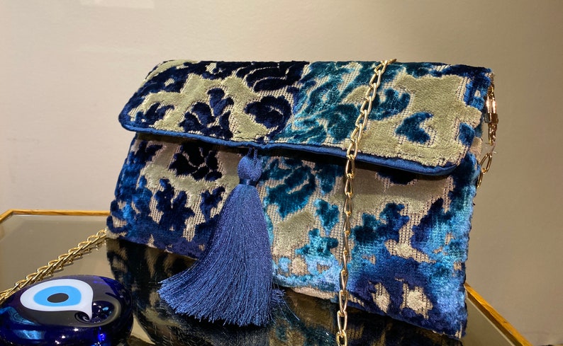 Op de afbeelding: Een blauwe en grijze velvet clutch met een gouden kettingband en een blauwe kwast. De tas heeft een bloemenpatroon.