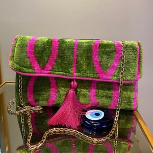 May include: A green and pink velvet clutch bag with a gold chain strap, a pink tassel, and a blue evil eye charm.