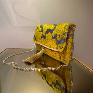 K&ouml;nnte beinhalten: Eine gelbe und blaue Clutch mit einem goldenen Kettenriemen und einem Quastenanh&auml;nger. Die Tasche liegt auf einer reflektierenden Oberfl&auml;che.