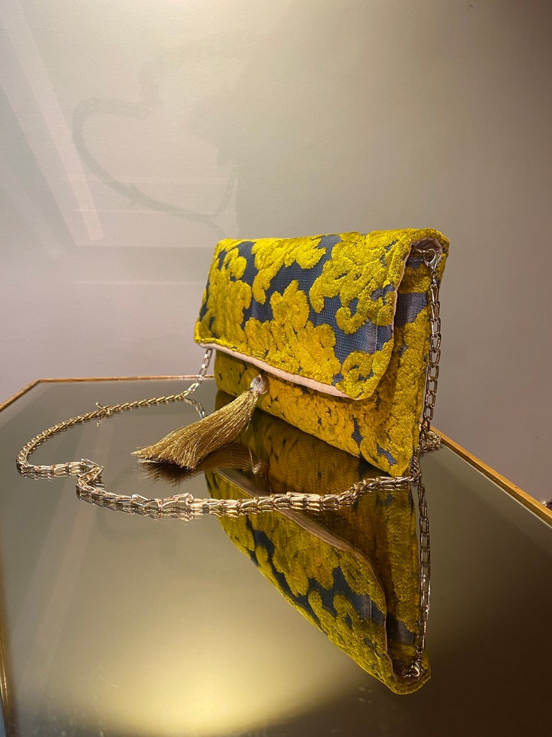 K&ouml;nnte beinhalten: Eine gelbe und blaue Clutch mit einem goldenen Kettenriemen und einem Quastenanh&auml;nger. Die Tasche liegt auf einer reflektierenden Oberfl&auml;che.
