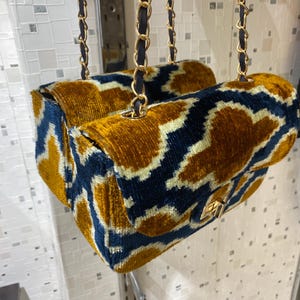 May include: Two velvet handbags with a gold and navy blue geometric pattern. The bags have gold chain straps with navy blue leather accents and gold clasps. The bags are hanging in front of a white wall.