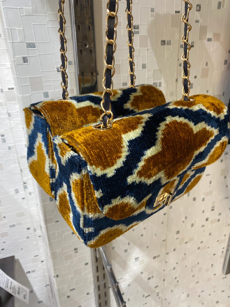 May include: Two velvet handbags with a gold and navy blue geometric pattern. The bags have gold chain straps with navy blue leather accents and gold clasps. The bags are hanging in front of a white wall.