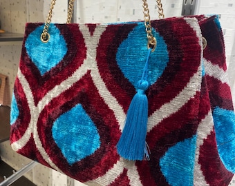 Könnte beinhalten: Gemusterte Handtasche mit goldfarbenem Kettenriemen. Die Tasche hat ein sich wiederholendes Design aus roten, weißen und türkisfarbenen Formen. Eine türkisfarbene Quaste hängt an der Seite. Die Tasche ist aus weichem, samtigem Material.