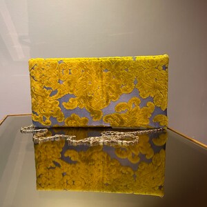 K&ouml;nnte beinhalten: Eine gelbe und blaue Clutch aus Samt mit einer goldenen Kettenkette. Die Clutch hat ein Blumenmuster und spiegelt sich in einem Spiegel.