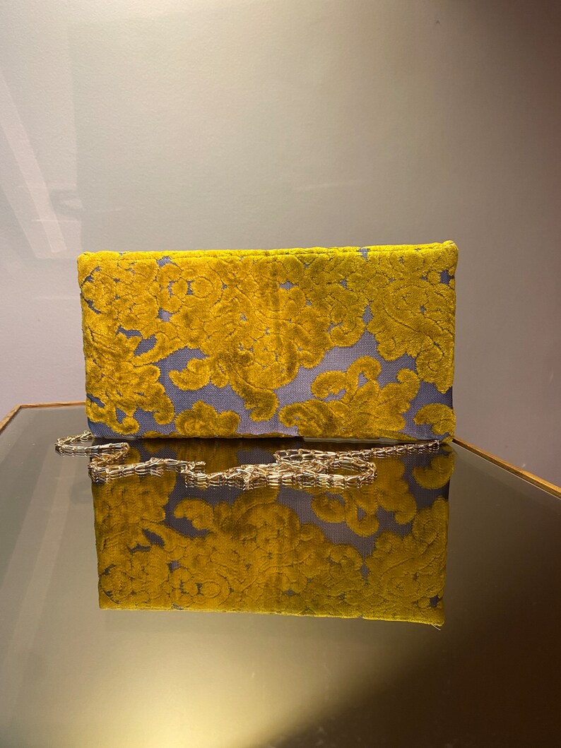 K&ouml;nnte beinhalten: Eine gelbe und blaue Clutch aus Samt mit einer goldenen Kettenkette. Die Clutch hat ein Blumenmuster und spiegelt sich in einem Spiegel.