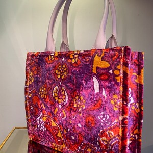 May include: A purple velvet tote bag with a floral pattern in shades of orange, pink, and white. The bag has a light purple leather handle.