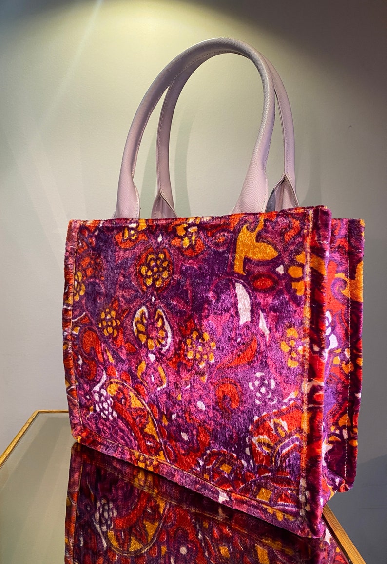 May include: A purple velvet tote bag with a floral pattern in shades of orange, pink, and white. The bag has a light purple leather handle.