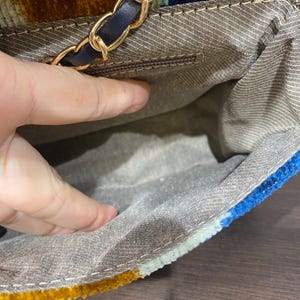May include: A colorful handbag with a gold chain strap and a navy blue leather accent. The bag's exterior features a textured pattern in shades of gold, blue, and cream. The interior is a light brown fabric.