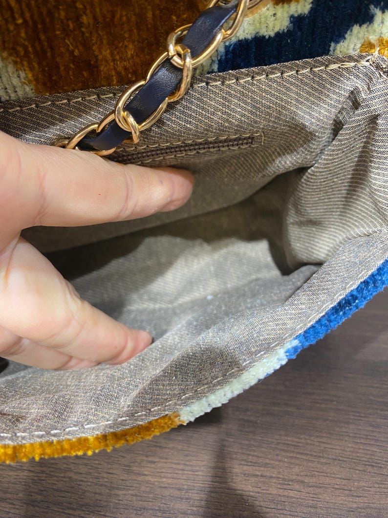 May include: A colorful handbag with a gold chain strap and a navy blue leather accent. The bag's exterior features a textured pattern in shades of gold, blue, and cream. The interior is a light brown fabric.