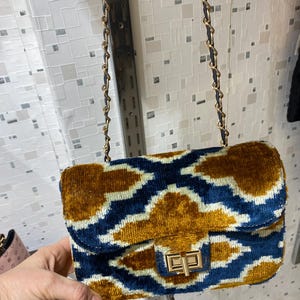 May include: A rectangular handbag with a gold chain strap. The bag features a blue and gold patterned design with a gold clasp. The bag is made of a soft, velvety material.