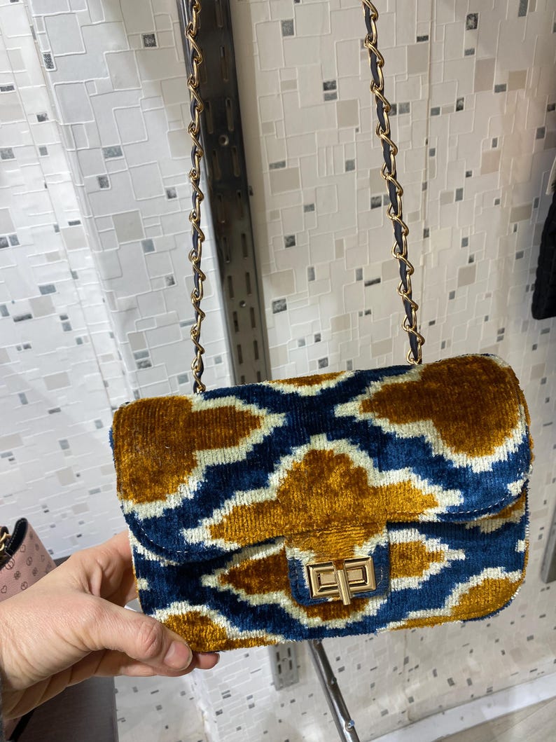 May include: A rectangular handbag with a gold chain strap. The bag features a blue and gold patterned design with a gold clasp. The bag is made of a soft, velvety material.