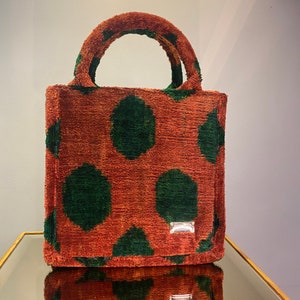 May include: A small, rectangular tote bag with a textured, rust-colored fabric featuring large, dark green polka dots. The bag has matching handles and a small, silver-toned rectangular tag on the front.