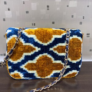 May include: A rectangular handbag with a gold chain strap. The bag features a velvet pattern in shades of blue, gold, and white. The chain strap is gold with a blue leather trim. The bag is a stylish accessory.