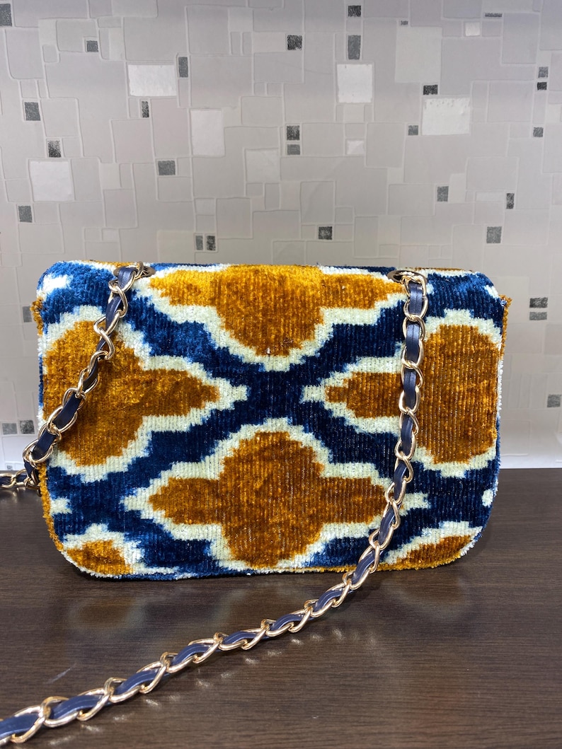 May include: A rectangular handbag with a gold chain strap. The bag features a velvet pattern in shades of blue, gold, and white. The chain strap is gold with a blue leather trim. The bag is a stylish accessory.