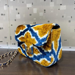May include: A velvet shoulder bag with a gold chain strap. The bag features a blue and gold geometric pattern and a gold clasp. The bag is sitting on a brown surface.