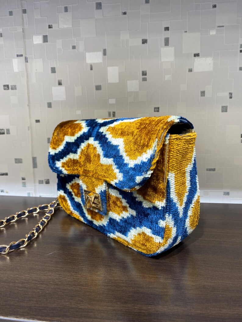 May include: A velvet shoulder bag with a gold chain strap. The bag features a blue and gold geometric pattern and a gold clasp. The bag is sitting on a brown surface.
