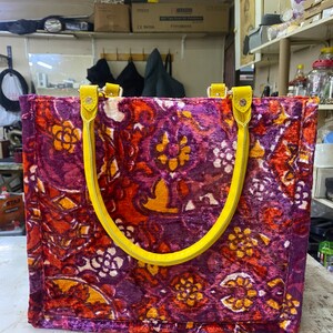 May include: A large tote bag with yellow handles. The bag is made of a purple, orange, and yellow fabric with a floral pattern.