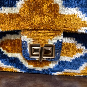 May include: Close-up of a handbag with a geometric pattern in blue, white, and gold. The bag has a textured, velvet-like appearance and a gold-colored clasp. The design features a repeating pattern of shapes.
