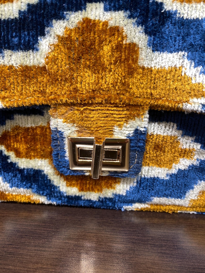 May include: Close-up of a handbag with a geometric pattern in blue, white, and gold. The bag has a textured, velvet-like appearance and a gold-colored clasp. The design features a repeating pattern of shapes.