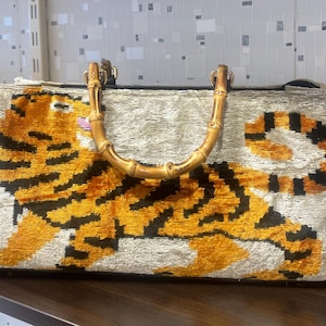 May include: A rectangular handbag with a tiger design in orange and black on a light gray background. The bag has a bamboo handle and a zipper closure. The bag is approximately 12 inches wide.