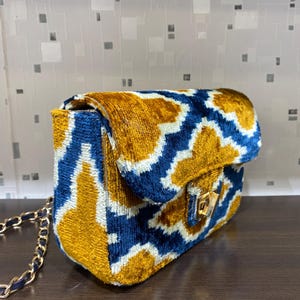 May include: A rectangular handbag with a gold chain strap. The bag features a geometric pattern in blue, gold, and white. The flap closure has a gold-colored clasp.