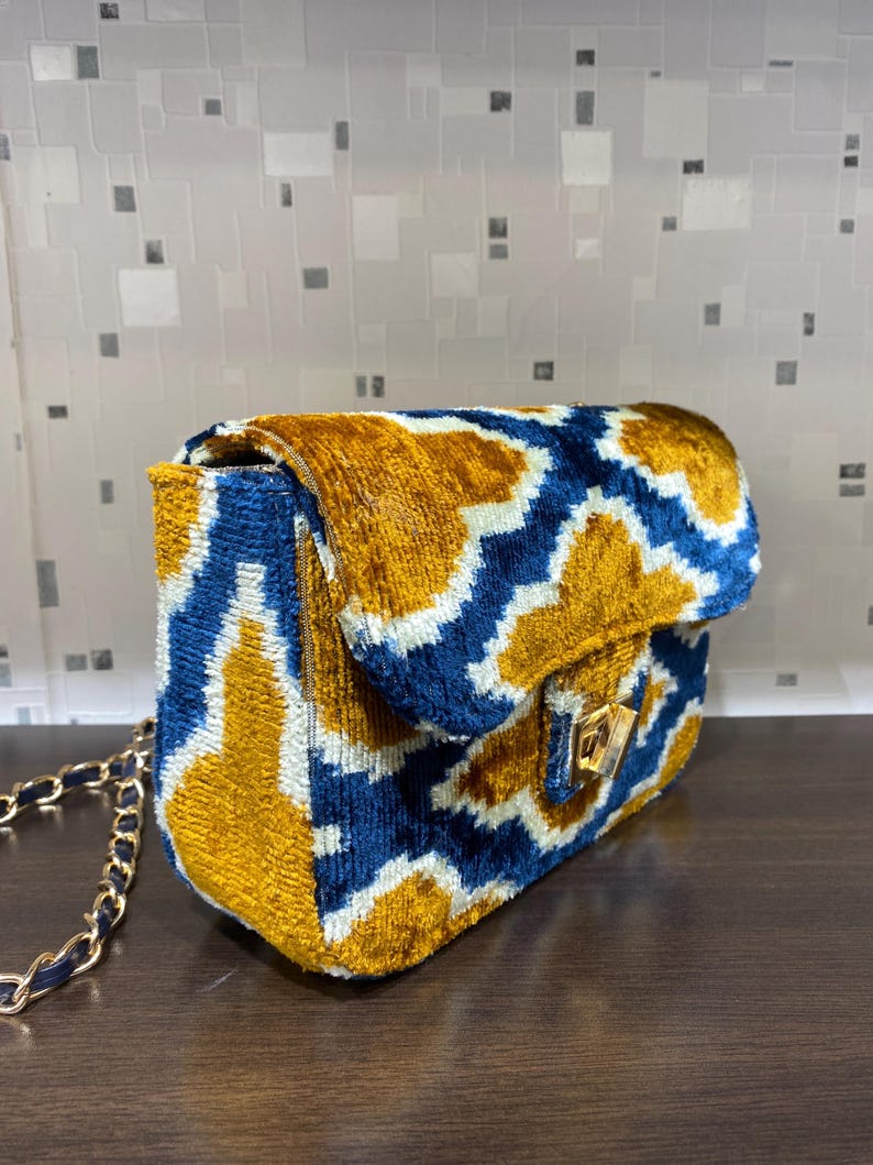 May include: A rectangular handbag with a gold chain strap. The bag features a geometric pattern in blue, gold, and white. The flap closure has a gold-colored clasp.