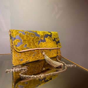 K&ouml;nnte beinhalten: Eine goldene Kettenhandtasche mit Quaste und Blumenmuster. Die Tasche ist aus weichem, samtigem Material.
