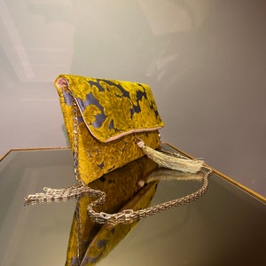 K&ouml;nnte beinhalten: Eine goldene Clutch aus Samt mit Blumenmuster und einer goldenen Kettenkette. Die Clutch hat einen Quasten-Anh&auml;nger.