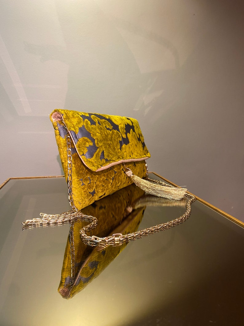 K&ouml;nnte beinhalten: Eine goldene Clutch aus Samt mit Blumenmuster und einer goldenen Kettenkette. Die Clutch hat einen Quasten-Anh&auml;nger.