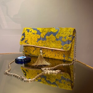 K&ouml;nnte beinhalten: Eine gelbe und blaue Clutch aus Samt mit einer goldenen Kettenkette und einem Quastenanh&auml;nger. Ein blauer b&ouml;ser Blick-Anh&auml;nger ist an der Kette befestigt.