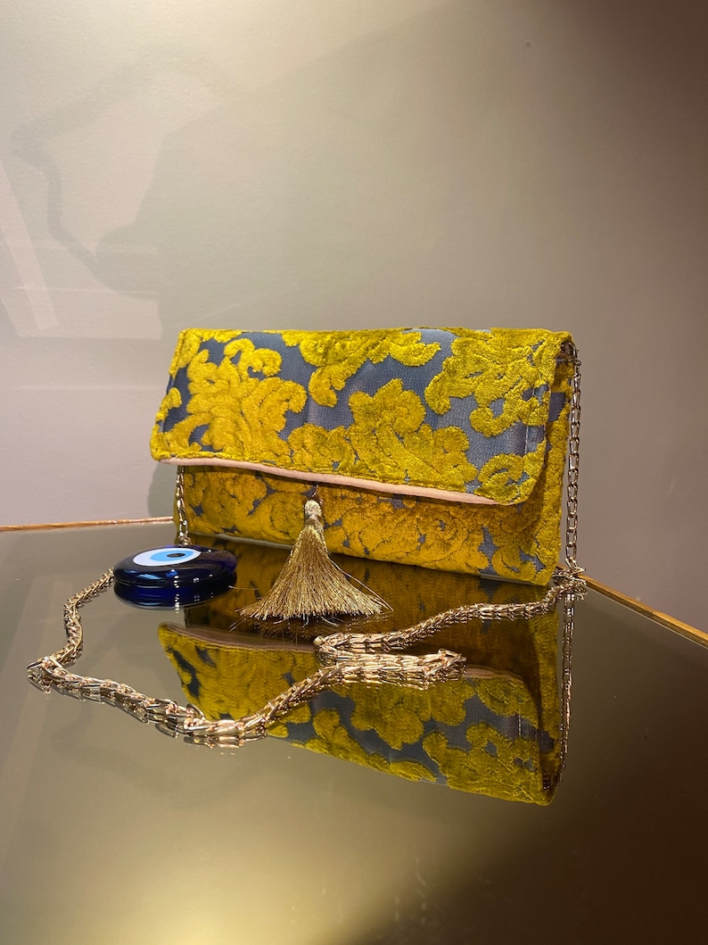 K&ouml;nnte beinhalten: Eine gelbe und blaue Clutch aus Samt mit einer goldenen Kettenkette und einem Quastenanh&auml;nger. Ein blauer b&ouml;ser Blick-Anh&auml;nger ist an der Kette befestigt.